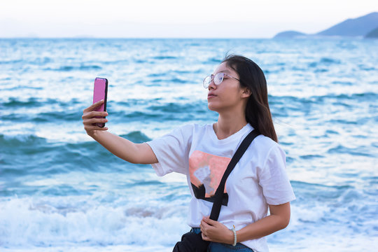 Young woman makes selfie on the beach on her smartphone. A girl with an Asian appearance stood on a berug against the background of the sea, a photograph on a mobile phone