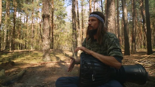 Young Caucasian hippie man playing a djembe drum in the summer coniferous forest