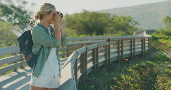 Young beautiful woman on vacation taking a picture outdoors with her vintage camera, happy attractive traveler woman taking a photograph of nature on a coastal boardwalk