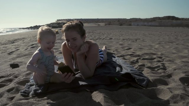 A young girl with her young child is resting on the ocean coast on a sandy beach. Mom shows her daughter how to use a smartphone. Technology and children. Vacation at sea in travel. New generation.