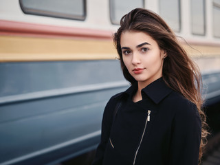 Young beautiful brown haired woman in black jacket blue skirt with gorgeous long legs in vintage beret emotionally posing on railway station train background with cinematic color grading