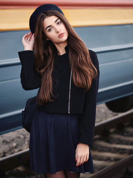 Young beautiful brown haired woman in black jacket blue skirt with gorgeous long legs in vintage beret emotionally posing on railway station train background with cinematic color grading