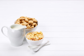 Corn flakes with milk in jar and spoons on white wooden table
