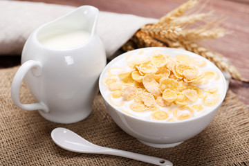 Corn flakes in bowl with milk and jar on brown wooden table