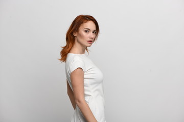 Beautiful redhead girl in a beige dress posing in front of the camera. Portrait on a neutral light background, space for text. Side view