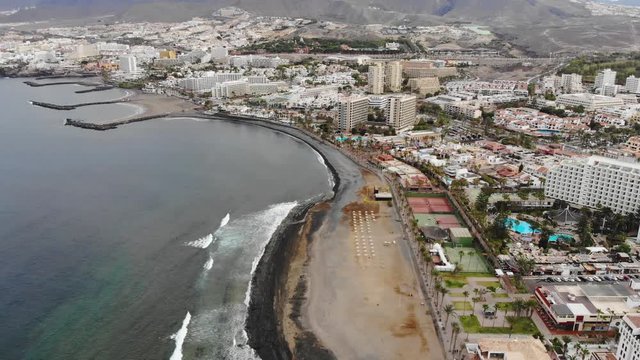 Aerial view of Las Americas city, Tenerife, Canary islands. Cinematic 4K footage with black volcanic rocky coast, Atlantic ocean, waves, city modern villas, quay with people, mountains. Bird eye shot