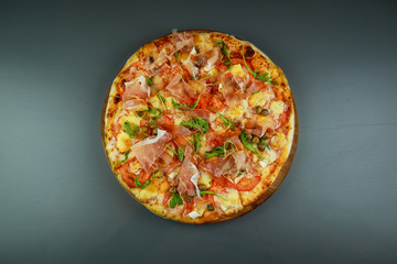 The pizza is prosciutto. Capers, tomatoes, arugula, mozzarella, brie cheese. Wooden plate. Gray background. Restaurant