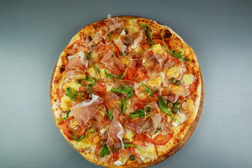 The pizza is prosciutto. Capers, tomatoes, arugula, mozzarella, brie cheese. Wooden plate. Gray background. Restaurant