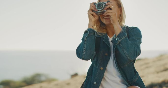 Happy attractive woman smiling and taking photographs with her vintage film camera outdoors by the ocean, young woman traveler on her dream adventure