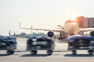 Luggage motion blurred trolley cart going fast delivering passenger baggage to modern plane on taxiway at airport on bright sunny day. Commercial aircraft on background at sunset or sunrise time