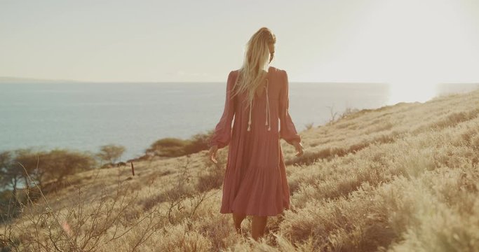 Beautiful blonde woman in a pink flowy dress walking through golden fields at sunset, fashion model photography