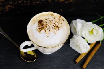 Cup of cappuccino with cinnamon and white flowers on dark background.