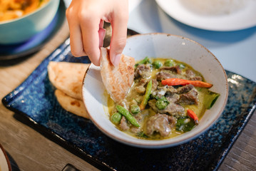 The hand of a woman holding a crispy roti, dip into the green curry