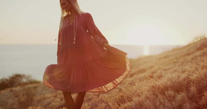 Happy carefree beautiful woman twirling her pink dress in the wind at sunset in a golden field by the sea, beautiful natural fashion model dancing at sunset