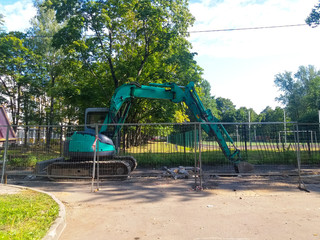 In the city yard, against the background of grass and trees behind a metal fence, a turquoise excavator digs up the roadbed. Russia, the city of St. Petersburg, August 2019.