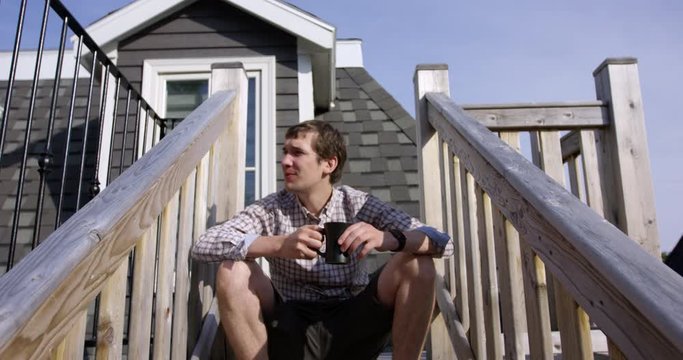 Young stylish man sitting on rooftop staircase enjoying morning coffee - slow tilt up