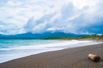 Black/ dark sand beach shore with heavy rain clouds above. Beautiful coastal landscape and low tide ocean water. Log/ driftwood on the seashore in Jamaica, an island in the tropical Caribbean sea.