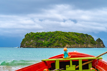 Fototapeta na wymiar Toy doll sitting in front colorful old wooden fishing boat docked by water on beautiful beach coast sea shore landscape on tropical Caribbean island. Holiday weekend summer vacation setting in Jamaica