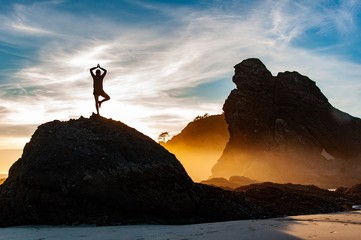 Young woman stands in tree pose among sea stacks at sunset