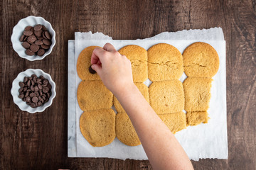 Two bowls of chocolate chips and batch of fresh cooked sugar cookies on parchment paper, woman’s hand putting chocolate on cookies