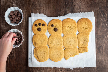 Two bowls of chocolate chips and batch of fresh cooked sugar cookies on parchment paper, woman’s hand putting chocolate on cookies