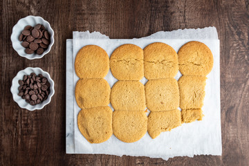 Two bowls of chocolate chips and batch of fresh cooked sugar cookies on parchment paper, on a wood table
