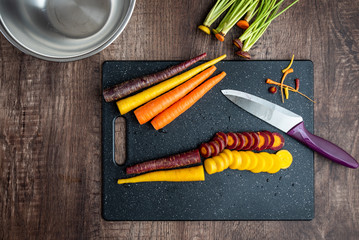 Sliced rainbow carrots on a black cutting board, chef knife, stainless-steel bowl, wood table