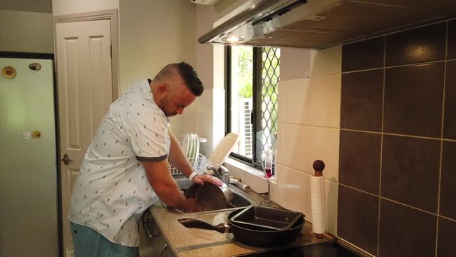 guy in the kitchen enjoying the view out the window whil ehe washes a large coffee pot in the sink before rinsng it and placing it on the stove top to dry
