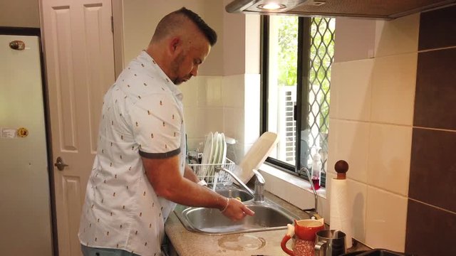 man at the sink in the kitchen washing the cutlery individually and rinsing and placing in the dish rack