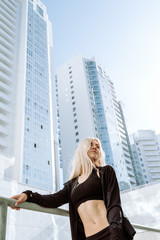 Fototapeta na wymiar Young adult blond female athlete in black sport clothes standing in front of tall city buildings, selective focus