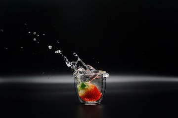 A strawberry drops into a glass of clear water causing water splash on the black background