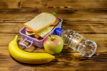 Lunch box with sandwiches, bottle of water, banana and apple on a wooden table