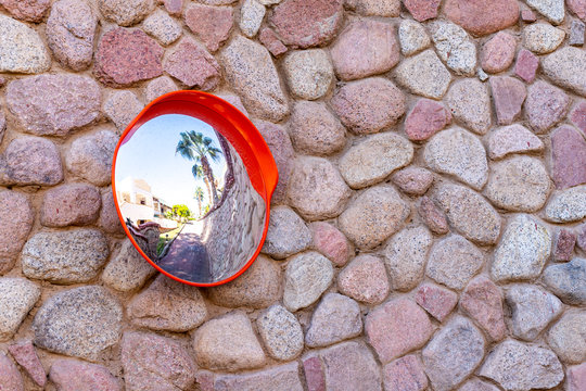 Convex Safety Mirror On Cobblestone Wall With Reflection Of Roadside View. A Classic Stone Wall And A Mirror For Viewing The Turn. A Mirror For Drivers To See What's Around The Corner.