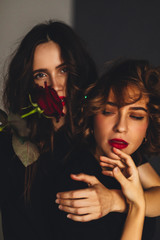 Image of a pretty young two women with bright red lipstick holding roses. Curly brunette hair woman wear black t-shirt. Image of a beautiful young cheery positive two women with bright red lipstick.