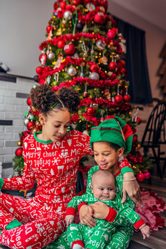 Siblings Sitting On The Floor Under The Tree Wearing Matching Christmas Holiday Pajamas.