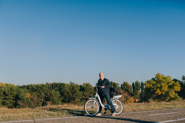 Obraz premium An elderly man, a pensioner rides an electric white bike on an asphalt road against the background of autumn nature. The concept of a happy old age. Fatigue and rest on the sidelines.