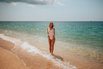 Beautiful girl in a white bodysuit walks along the shore of the azure sea