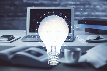 Computer on desktop in office with bulb icon hologram. Multi exposure. Concept of idea.