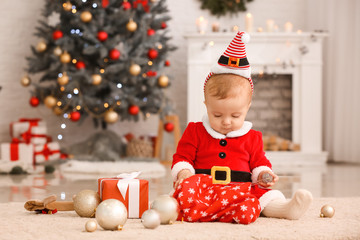 Cute little baby in Santa Claus costume and with gifts in room decorated for Christmas