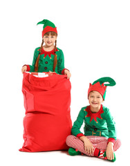 Little children in costume of elf and with Santa Claus bag full of gifts on white background