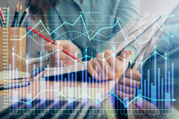 A woman hands writing information about stock market in notepad. Forex chart holograms in front. Concept of research. Multi exposure