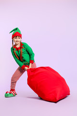 Little girl in costume of elf and with big Santa Claus bag on color background