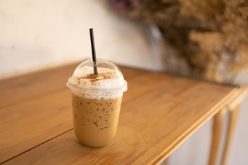 Tasty drinking, a cup of ice cappuccino coffee decorated with white milk froth in a tall plastic glass on brown wooden table, blurred died flower and white background