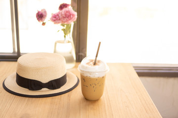 Tasty drinking, cup of ice cappuccino coffee decorated with white milk froth in a tall plastic glass on brown wooden table, beside a hat and pink roses in a vase, window on background