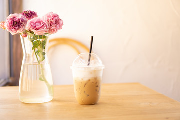 Tasty drinking, cup of ice cappuccino coffee decorated with white milk froth in a tall plastic glass on brown wooden table, near roses in a glass vase with water, blurred white background