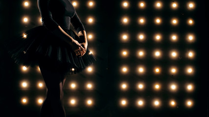 Silhouette of ballerina in black tutu is dancing ballet in the dark studio with the lights on the background. © alexeg84