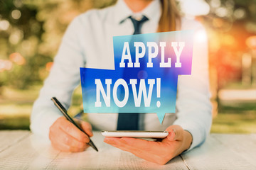 Word writing text Apply Now. Business photo showcasing request something officially in writing or by sending in form Female business person sitting by table and holding mobile phone