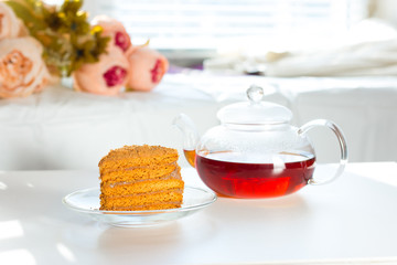 Breakfast concept. Black tea in a glass cup and a teapot on a white table. A slice of honey cake on a transparent saucer. Breakfast for a beloved woman.
