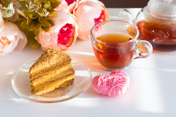 Black tea in a glass cup on a white table. A slice of honey cake on a transparent saucer. A bouquet of pink flowers and a gift with a red bow. International Women's Day. Valentine's Day.