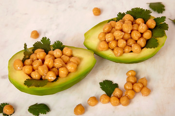 Half cut avocado with boiled chickpeas and parsley inside, vegan diet meal on marble background. Healthy food concept, indoors.